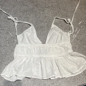 Vit ärmlös topp med volang - Säljer en söt helt oanvänd vit topp i storlek S. Den är ärmlös med smala axelband som knyts på axlarna. Toppen har en volangdetalj vid midjan och är gjord i ett lätt och luftigt material, perfekt för sommaren. Passar bra till både vardag och fest! 