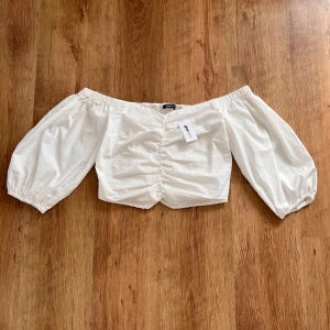 Vit off-shoulder topp från Gina Tricot - Säljer en superfin vit off-shoulder topp från Gina Tricot. Toppen är i bomull och har puffiga långärmar med elastiska ärmslut. Framsidan har en snygg rynkad detalj och baksidan är smockad för en bekväm passform. Perfekt för sommarens alla fester! 🌸
