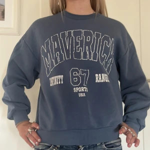 Blå sweatshirt med tryck - Säljer en blå sweatshirt med tryck på framsidan. Texten säger 'MAVERICK' och 'UNITY RANGER 67 SPORTS USA'. Tröjan är långärmad och har en normal passform. Perfekt för en avslappnad stil eller för att hålla sig varm under kyliga dagar. 