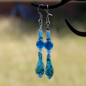 Blå örhängen med glaspärlor - Säljer ett par superfina blå örhängen med glaspärlor. De har en vacker droppformad design med olika nyanser av blått och små glittriga detaljer inuti pärlorna. Den droppformade pärlan lyser i mörkret!