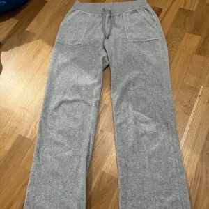Grå mjukisbyxor från Juicy Couture - Säljer ett par superbekväma grå mjukisbyxor från Juicy Couture i storlek S. Byxorna har en hög midja med snörning och två fickor fram samt två fickor bak med knappar. Perfekta för en mysig dag hemma eller en avslappnad dag ute. 🩶