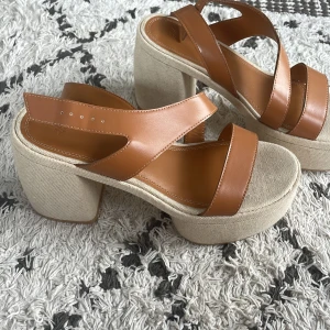 Bruna sandaler med klack - Säljer ett par snygga bruna sandaler med klack. De har en bekväm och stabil klack i beige tygmaterial och remmar i brunt skinn. liten fläck på ena skon, men syns ej vid användning då de är vid tån.