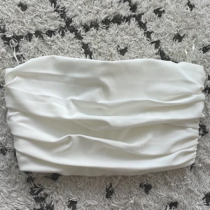 Vit tubtopp från Zara - Säljer en snygg vit tubtopp från Zara. Toppen är i ett mjukt och stretchigt material som sitter bekvämt på kroppen. Perfekt för sommaren eller en festkväll! Toppen är i nyskick och har en stilren design med rynkade detaljer. 