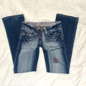 Blå jeans med slitningar - Säljer ett par utsvängda blåa lågmidjade bootcut jeans. Jeansen har coola detaljer som röda sömmar, strl S/36. Jeansen är också ganska stretchiga i midjan❤️