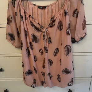 Rosa blus med fjädermönster - Säljer en söt rosa blus från H&M med ett fint fjädermönster i svart och rosa. Blusen har trekvartsärmar och en knytning framtill vid halsen. Perfekt för vår och sommar! 🌸