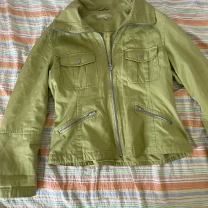 Grön jacka från KappAhl - Säljer en snygg grön jacka från KappAhl i storlek L. Jackan har en dragkedja framtill och två bröstfickor med knappar samt två snedställda fickor med dragkedja. Perfekt för vår och höst!