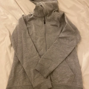 Grå hoodie från Kappa - Säljer en stilren grå hoodie från Kappa med dragkedja och huva. Perfekt för en avslappnad look. Den har långa ärmar och praktiska fickor framtill. Passar bra till både vardag och träning.