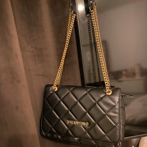 Svart axelväska Valentino - Snygg svart axelväska från Valentino i quiltad design med guldfärgade kedjedetaljer. Endast använd 5 ggr. Defekt på undersidan, se bild 2, dock inget som syns tydligt men värt att nämna. Köpt på zalando för ca 1500kr. Dust bag medföljes.💝