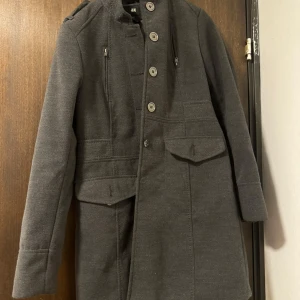 Grå kappa från H&M - Snygg grå kappa från H&M med knappar framtill och dekorativa knappar på ryggen. Kappan har en klassisk design med långa ärmar och en stilren look. Perfekt för kyligare dagar.