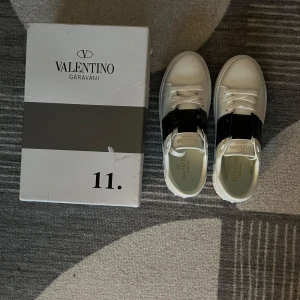 Valentino Garavani sneakers - Snygga Valentino Garavani sneakers i vitt med en bred svart rem över ovansidan. Skorna har klassisk snörning och en stilren design som passar perfekt för en trendig look. Grisch asf. Aldrig använt.