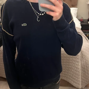 Mörkblå tröja från Lacoste - Säljer en stilren mörkblå tröja från Lacoste med deras fina logga på bröstet. Tröjan har långa ärmar och kontrasterande detaljer längs axlarna💗Säljer den då jag inte använder den länge❤️tröjan är i storlek 36 och passar jättebra för mig som är 167❤️tröjan har inga tecken på användning och nypris är 1355