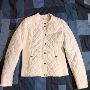 Croppad Vit vintage  quiltad jacka med dragkedja och knappar - Snygg vit quiltad jacka med både dragkedja och knappar framtill. Jackan har en stilren design med långa ärmar och ribbade sidopaneler för extra komfort. Perfekt för kyligare dagar. Tyvärr så har sidopanelerna blivit noppriga med året men inget som märks under användning eller syns. Dm vid frågor