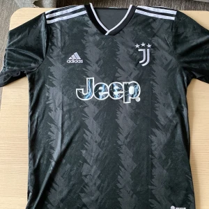 Svart Juventus fotbollströja från Adidas - Snygg svart Juventus fotbollströja från Adidas med korta ärmar och ett diskret mönster. Tröjan har vita detaljer och Juventus-loggan på bröstet samt Jeep som sponsor. Perfekt för alla fotbollsfans!