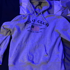 Grå hoodie från Polo Club - Säljer en grå hoodie från Polo Club Saint Vincent med tryck framtill och en amerikansk flagga. Tröjan har en klassisk känguruficka och justerbar huva med dragsko. Perfekt för en avslappnad stil.