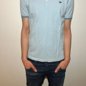 Ljusblå pikétröja från Emporio Armani - Snygg ljusblå pikétröja från Emporio Armani med korta ärmar och diskret logga på bröstet. Tröjan har en klassisk krage och knappar framtill. Perfekt för en stilren look.