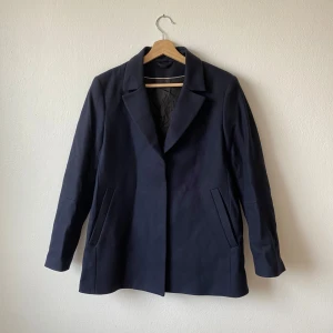 Mörkblå kort kappa från J.Lindeberg - Elegant mörkblå kappa från J.Lindeberg med klassisk design. Kappan har långa ärmar och en stilren krage. Perfekt för både höst och vår, med en tidlös look som passar till många tillfällen. Modell: Annie Short Coat - Artnr:G094