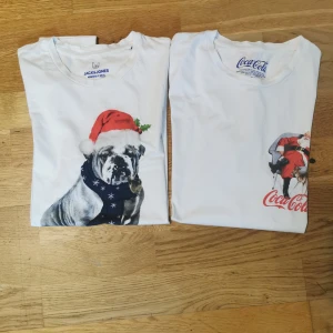 Två vita jul-t-shirts från Jack & Jones och Coca-Cola - Säljer två vita t-shirts med julmotiv. Den ena från Jack & Jones har en bulldog med tomteluva och texten 'Deck Me Out'. Den andra från Coca-Cola har en bild av jultomten med Coca-Cola-loggan. Perfekta för julfirandet!