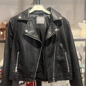 Svart skinnjacka från Lindex - Snygg svart skinnjacka från Lindex med dragkedja och knappar. Klassisk bikerstil med långa ärmar och stilrena detaljer. Perfekt för en cool look.