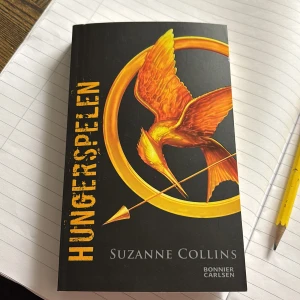 Hungerspelen - Säljer boken 'Hungerspelen' av Suzanne Collins, utgiven av Bonnier Carlsen. Omslaget är svart med en gyllene fågel och pilmotiv. Perfekt för dig som gillar spännande dystopiska berättelser.