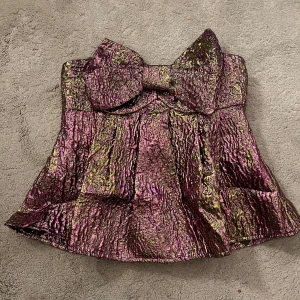 Glittrig korsettopp med rosett - Snygg glittrig korsettopp i lila och guld med en stor rosett framtill. Perfekt för en festlig kväll! Toppen har en strukturerad design och är axelbandslös. Stolek XS, helt oanvänd. 