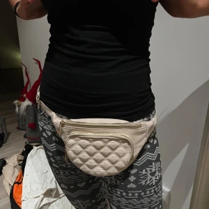 Beige quiltad magväska - Snygg beige magväska med quiltat mönster och dragkedja. Perfekt för att hålla dina saker säkra och nära till hands. Stilren och praktisk design.