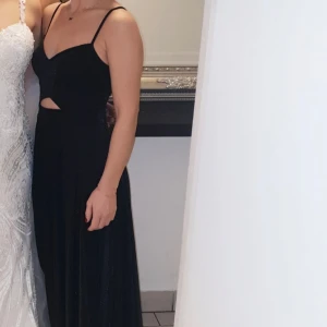Svart långklänning med cut-out detaljer - Elegant svart långklänning med smala axelband och stiliga cut-out detaljer i midjan. Perfekt för en festlig kväll. Lite glittrig i tyget men ingenting som ser för mycket ut. Går att justera med banden i ryggen. Passar 34-36