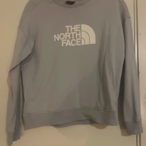 Blå  sweatshirt från The North Face - Säljer en stilren grå sweatshirt från The North Face med deras ikoniska logga i vitt på framsidan. Tröjan har långa ärmar och en bekväm passform, perfekt för kyligare dagar.