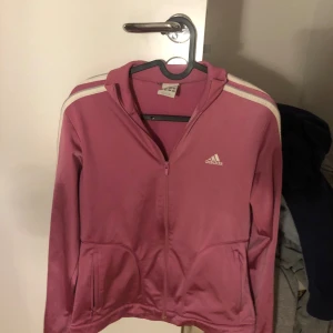 Rosa zip up från Adidas - Säljer en rosa träningsjacka från Adidas med dragkedja och vita ränder på axlarna. Jackan har långa ärmar och två fickor framtill. Perfekt för träning eller en avslappnad stil.