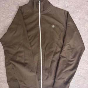Brun track jacket från Fred Perry - Säljer en snygg brun tracktop från Fred Perry med vit dragkedja och broderad logga på bröstet.  Skick 10/10