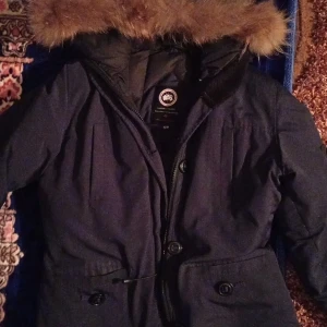 Mörkblå parkajacka från Canada Goose - Säljer en mörkblå parkajacka från Canada Goose med pälsfodrad huva. Jackan har knappar och dragkedja framtill samt en ikonisk logga på ärmen. Perfekt för kalla vinterdagar.