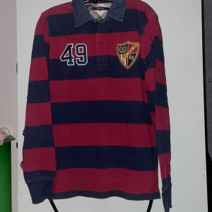 Randig rugbytröja från GANT - Säljer en randig rugbytröja från GANT i rött och blått med nummer 49 och ett broderat emblem på bröstet. Tröjan har en krage i denim och långa ärmar. Perfekt för en sportig look!