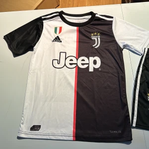 Juventus fotbollströja och shorts - Snygg Juventus fotbollströja med korta ärmar och matchande shorts. Tröjan är svart och vit med en röd rand och har Juventus-loggan samt 'Ronaldo 7' på baksidan. Shortsen är svarta med vita ränder och Juventus-loggan. Ganska liten i storleken. Perfekt för fotbollsfans!