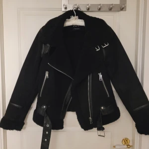 Svart jacka med dragkedjor och bälte - Snygg svart jacka med flera dragkedjor och bältesdetaljer. Jackan har en cool design med långa ärmar och en fluffig krage. Är i bra skick.<3