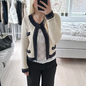 Beige och svart kofta med stickat mönster - Snygg beige kofta med svarta detaljer och stickat mönster. Koftan har långa ärmar och dekorativa knappar. Perfekt för en stilren look. Den är i storlek M från Lindex. Finns tecken på användning. Glittrig.