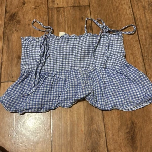 Blå- och vitrutigt linne från H&M - Charmigt blå- och vitrutigt linne från H&M med smockdetaljer och knytband vid axlarna. Perfekt för en somrig look med sin luftiga design och volangkant.