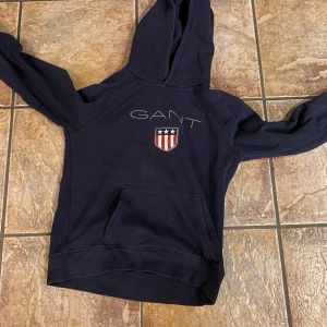 Mörkblå hoodie från GANT - Säljer en snygg mörkblå hoodie från GANT med broderad logga och emblem på bröstet. Perfekt för en avslappnad stil med en stor ficka framtill och bekväm huva. Tröjan är 158/164