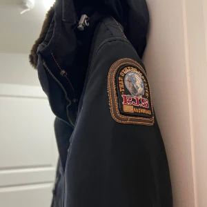 Svart parkajacka från Parajumpers - Säljer en svart parkajacka från Parajumpers med pälsfodrad huva och dragkedja. Jackan har flera praktiska fickor och en snygg patch på ärmen. Perfekt för kalla dagar.