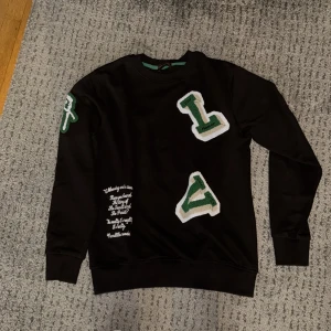 Svart sweatshirt från Louis Vuitton - Snygg svart sweatshirt med gröna och vita applikationer i form av bokstäver. Tröjan har långa ärmar och en rund halsringning. Perfekt för en avslappnad stil. 