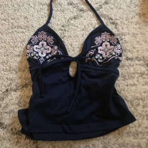 Svart broderad halterneck topp - Snygg svart halterneck topp med vackra broderier i rosa och grönt. Toppen har en knytning framtill och en liten öppning under bysten. Perfekt för en somrig look!