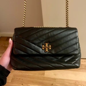 Svart quiltad axelväska med kedja - Snygg svart axelväska från Tory Burch i quiltat skinn med guldfärgad kedja. Väskan har ett elegant mönster och en logotyp på framsidan. Perfekt för att ge din outfit en lyxig touch.