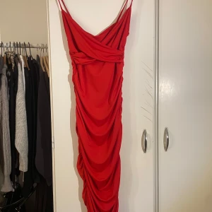 Röd omlottklänning - Snygg röd omlottklänning med smala axelband och draperad design. Perfekt för en kväll ute med v-ringad hals och figurnära passform. Klänningen har en elegant rynkad detalj längs hela längden som ger en smickrande siluett.