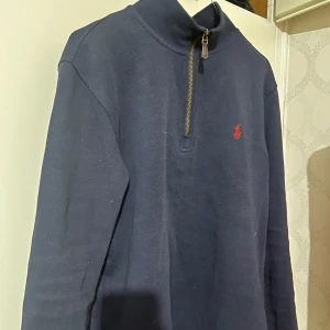 Mörkblå tröja från Polo Ralph Lauren - Säljer en stilren mörkblå tröja från Polo Ralph Lauren med dragkedja och det klassiska röda logotypbroderiet på bröstet. Perfekt för en avslappnad men ändå snygg look. Tröjan har långa ärmar och en hög krage.