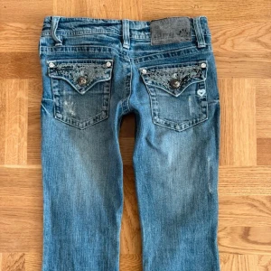Blå jeans med dekorativa bakfickor - Snygga ljus blå jeans med bootcut-stil och låg midja. Bakfickorna är dekorerade med glittriga detaljer och knappar. 