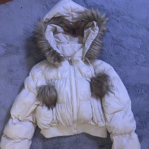 Beige pufferjacket - Säljer en beige pufferjacka med fuskpälsdetaljer på huvan och stora bollar framtill. Jackan är croppad och har dragkedja. Pris kan diskuteras. Storlek XS