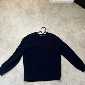 Mörkblå tröja från Ralph Lauren - Säljer en stilren mörkblå tröja från Ralph Lauren med det klassiska röda logotypen på bröstet. Tröjan har långa ärmar och ribbstickade muddar vid ärmslut och nederkant. Perfekt för en avslappnad och snygg look.