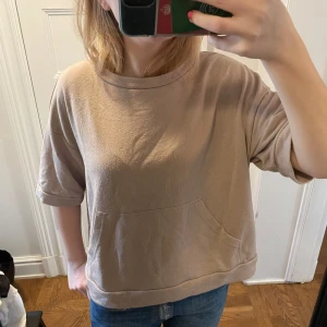 Beige tröja med ficka - Säljer en beige tröja med korta ärmar och en praktisk ficka framtill. Tröjan har en avslappnad passform och är perfekt för en casual look. Den är gjord i ett mjukt material som känns skönt mot huden.
