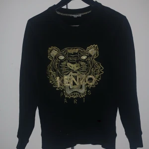 Svart tröja från Kenzo med tigerbroderi - Snygg svart tröja från Kenzo med ett riktigt schyst guldigt tigerbroderi på framsidan. Perfekt för vardan men även finare tillfällen! Säljer nu pga att den tyvärr har blivit för liten. Fint skick förutom små två små märken som inte märks. Tveka inte att höra av dig vid intresse!
