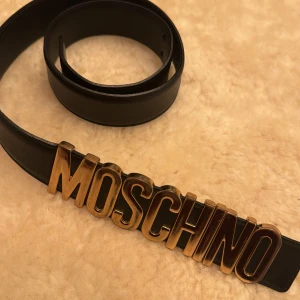 Svart skärp från Moschino med guld  - Snyggt svart skärp från Moschino i äkta skinn med en iögonfallande guldfärgad logotyp framtill. Tillverkat i Italien, perfekt för att ge en lyxig touch till din outfit. Strl 38. Har tyvärr ej kvitto men köp i London (äkta). Nypris ca 3000kr säljer för 600kr. Gott skick med någon skråma.