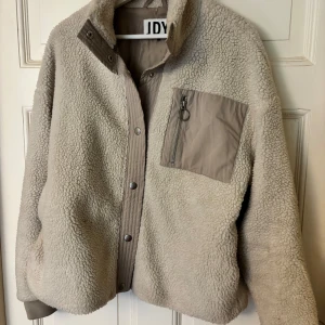 Beige teddyjacka från JDY - Mysig beige teddyjacka från JDY med dragkedja och knappar framtill. Jackan har en praktisk bröstficka med dragkedja i kontrasterande material. Perfekt för kyliga dagar.