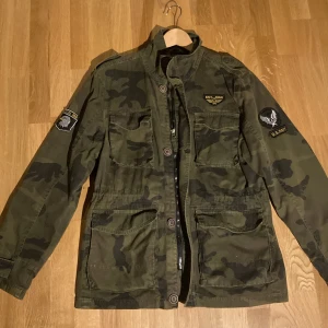 Millitär jacka - Säljer en grön camouflagejacka från Tokyo Laundry med coola patchar och knappar. Jackan har en dragkedja och knappar framtill samt flera praktiska fickor. Perfekt för en avslappnad stil. Perfekt för våren eller de lite varmare vinterdagarna.